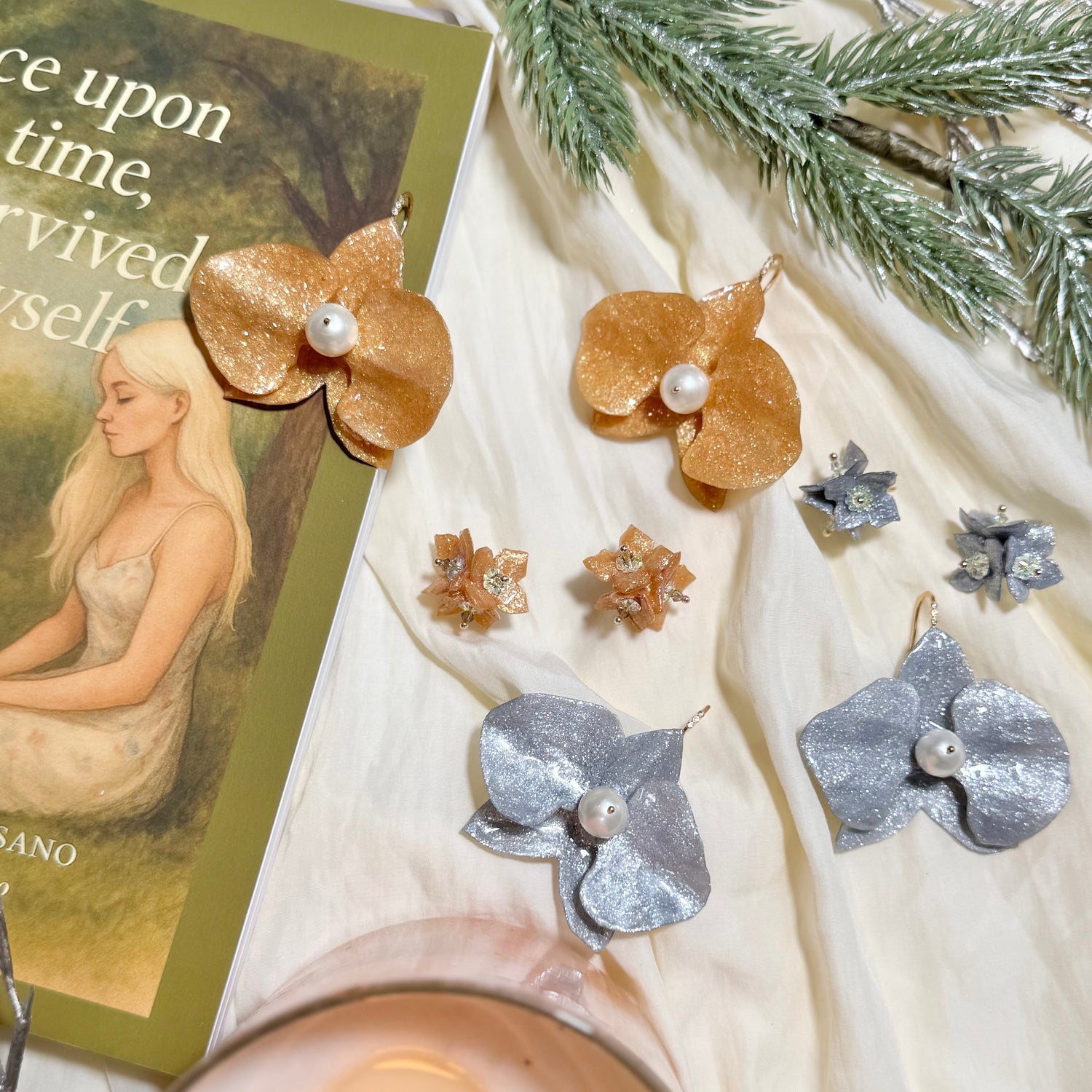 Pair of bronze flower-shaped earrings with pearl centers on a white fabric background with a candle and book.
