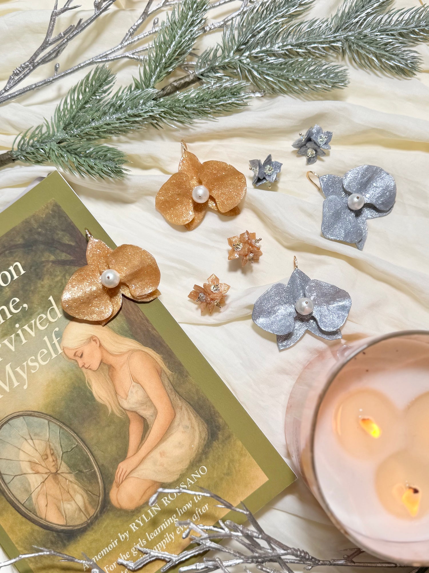 Gold and silver earrings on a textured surface with candles and decorative branches.